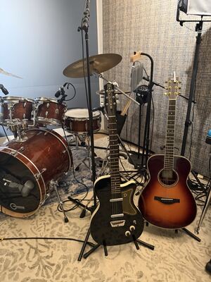 Item 10: black Danelectro single-cutaway electric guitar with a tan D pickguard, on a stand next to a drum kit